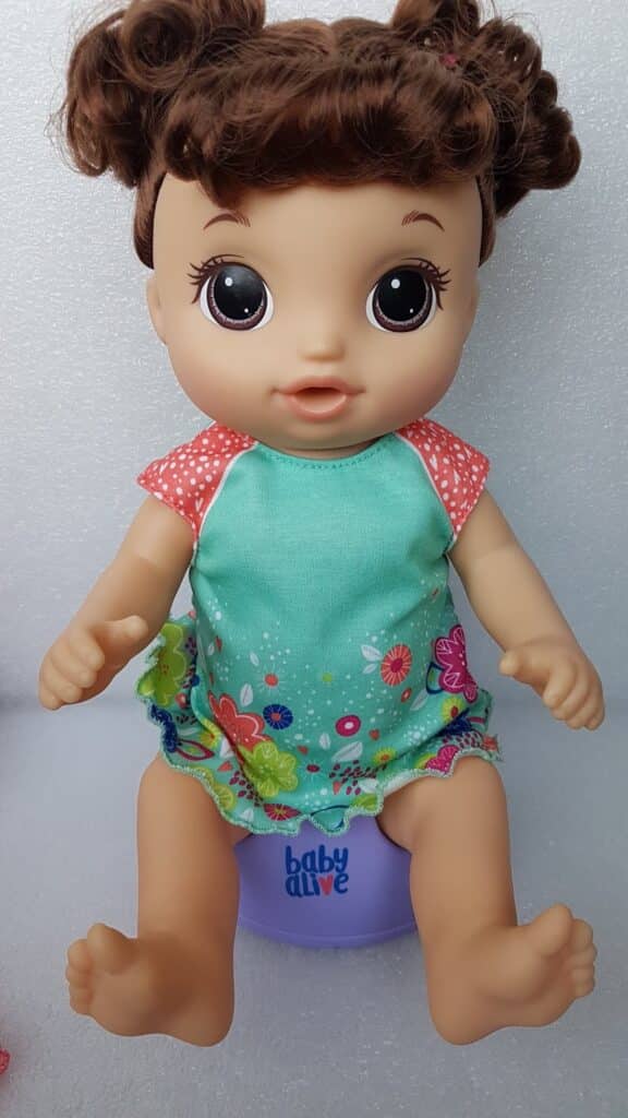 A Baby Alive doll with brown hair in bunches, wide brown eyes, and light skin sits on a purple potty labelled "baby alive." The doll wears a turquoise dress with floral patterns and pink spotty sleeves, and has an open-mouthed expression.