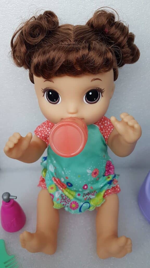 A baby doll with curly brown hair in bunches, big brown eyes, and wearing a colourful floral outfit sits upright. The doll holds a pink bottle to its mouth, as if drinking, with toys and accessories placed around it on a white background.