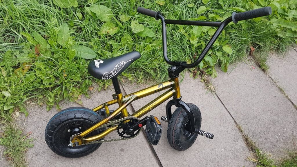 A small gold BMX bike with thick black tyres, black handlebars, and a black seat decorated with a white graphic, is parked on a concrete path beside tall green grass and leafy plants.