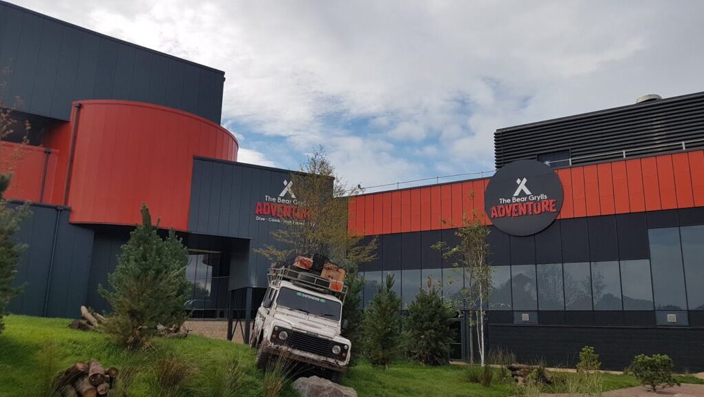 A modern building with dark grey and orange curved panels displays signs reading "The Bear Grylls Adventure." In front, an off-road vehicle loaded with gear is angled on a grassy slope, surrounded by small trees and rocks under a partly cloudy sky.