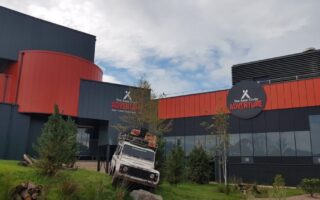 A modern building with dark grey and orange curved panels displays signs reading "The Bear Grylls Adventure." In front, an off-road vehicle loaded with gear is angled on a grassy slope, surrounded by small trees and rocks under a partly cloudy sky.