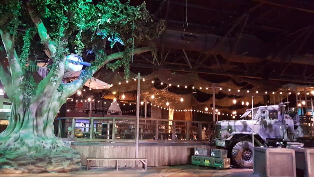 A large artificial tree with green lighting stands indoors beside a camouflaged military lorry. Fairy lights hang above a wooden-railed area with tables and chairs. The ceiling is exposed, and the scene has a rustic, adventure-themed atmosphere.