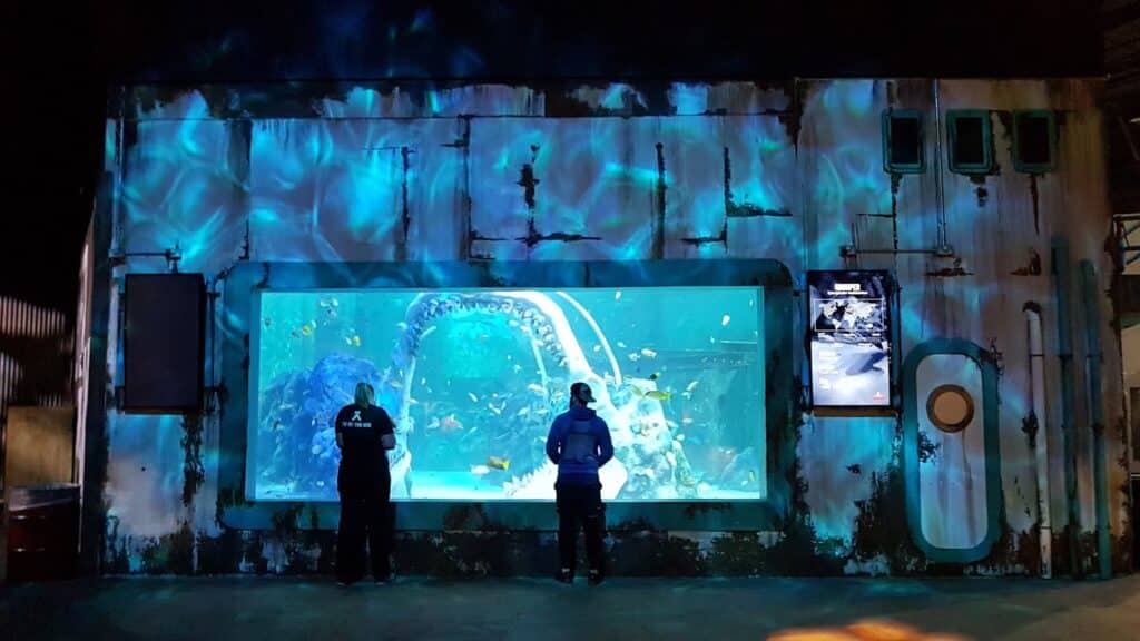 Two people stand in front of a large aquarium with blue lighting. The tank features a giant shark jaw model, colourful fish, and underwater-themed decor. The wall around the tank is weathered and industrial, with a door and screens displaying information nearby.
