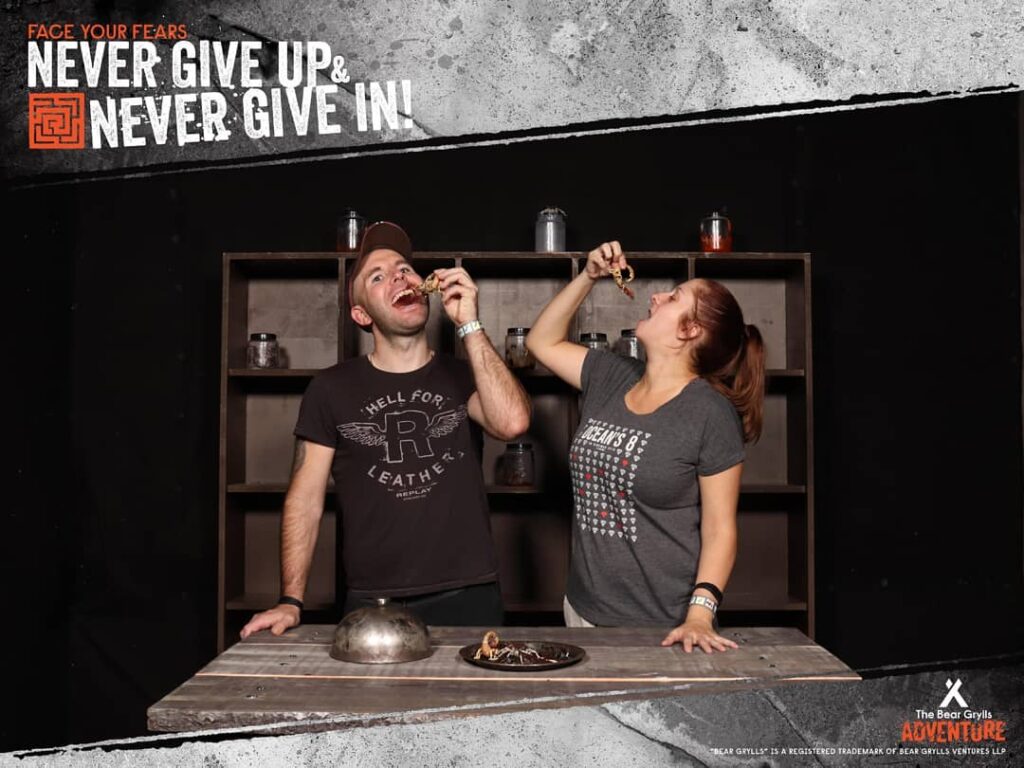 Two people stand in front of a shelf eating insects, one man and one woman, both wearing casual T-shirts and wristbands. The table holds a bowl and insects. Text above reads “NEVER GIVE UP & NEVER GIVE IN! Face your fears.” The atmosphere is adventurous.