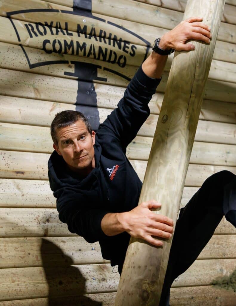 A man in a black hoodie climbs a large wooden pole indoors. Behind him, a log wall displays the words "ROYAL MARINES COMMANDO" in black text. The man looks determined, gripping the pole firmly with both hands.