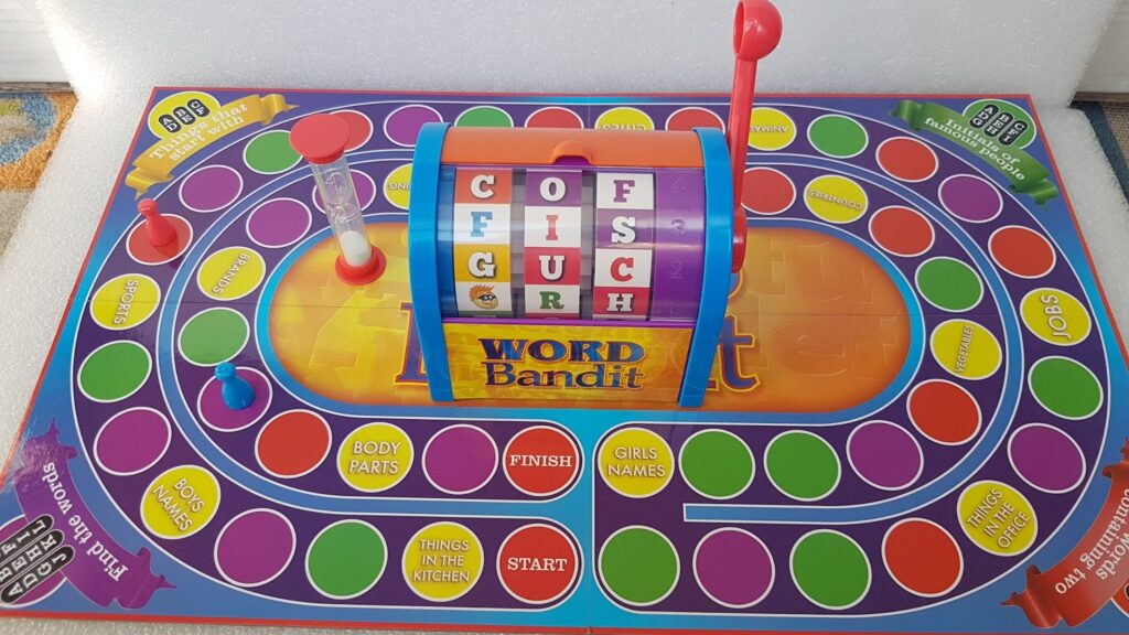 A colourful Word Bandit board game with a central fruit-machine style spinner showing letters. The board has a winding path of colourful circles, labelled categories like “BODY PARTS” and “THINGS IN THE KITCHEN”, and an hourglass on the left.