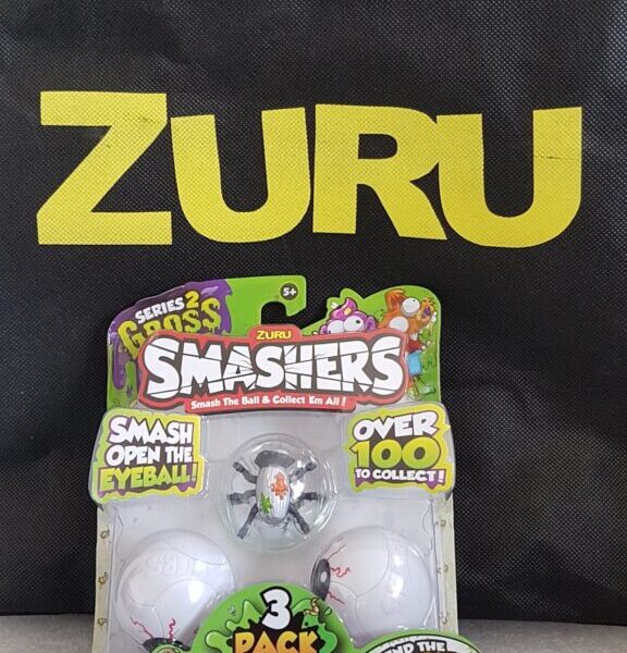 A ZURU-branded bag stands upright behind a packaged ZURU Smashers toy. The packaging features three ball-shaped collectables decorated as eyeballs and colourful cartoon graphics, with text promoting the toy series and collectable features.