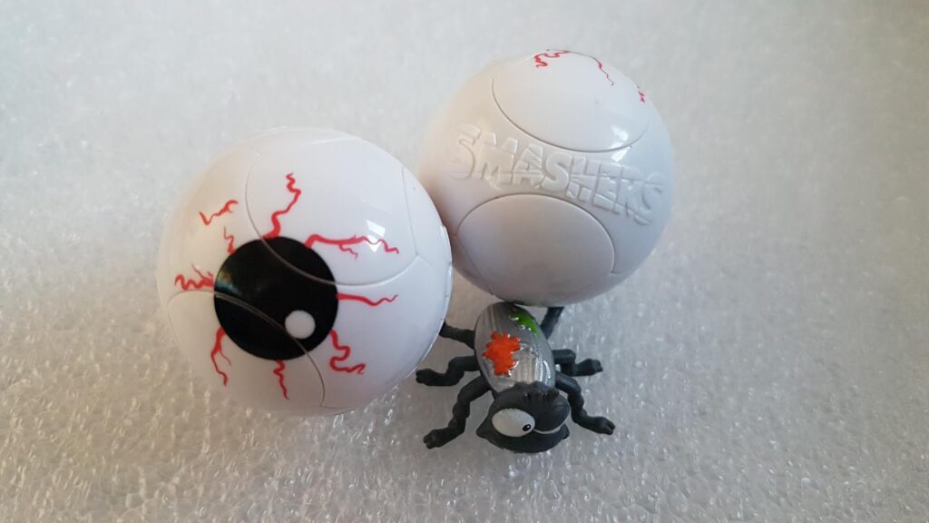 Two white plastic toy eyeballs with red veins, one fully closed and one partially open to reveal black and red details. A small toy spider with grey body, green accents, and orange spot is next to them. The background is textured white foam.