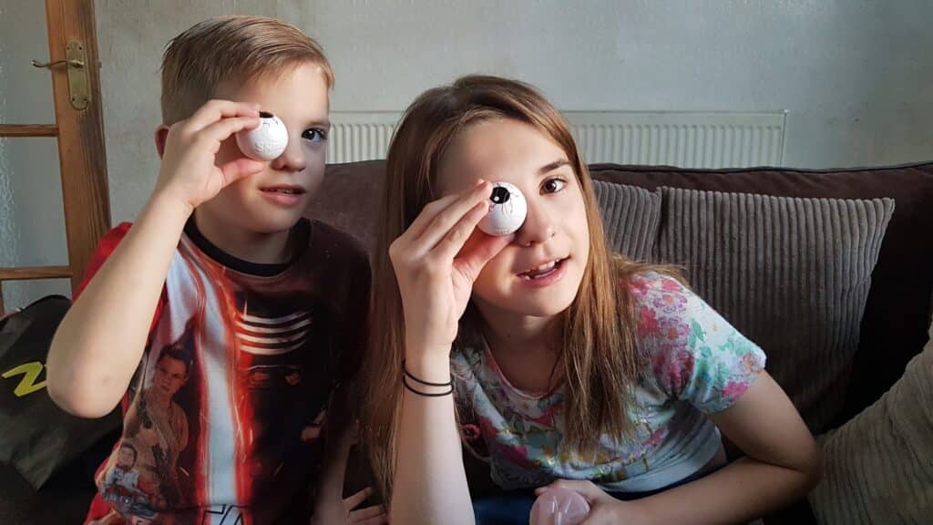 A young boy and girl sit on a sofa holding small white balls over one eye, pretending they are eyeballs. The boy wears a Star Wars shirt and the girl has a floral t-shirt. Both look amused, and the background shows a radiator and cushions.