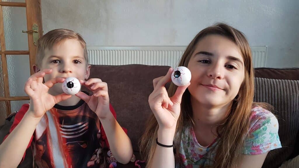 A boy and a girl sit on a brown sofa, smiling and each holding up a fake eyeball with red veins. The boy wears a dark shirt with a print, and the girl wears a floral top. A white radiator and wooden door are in the background.
