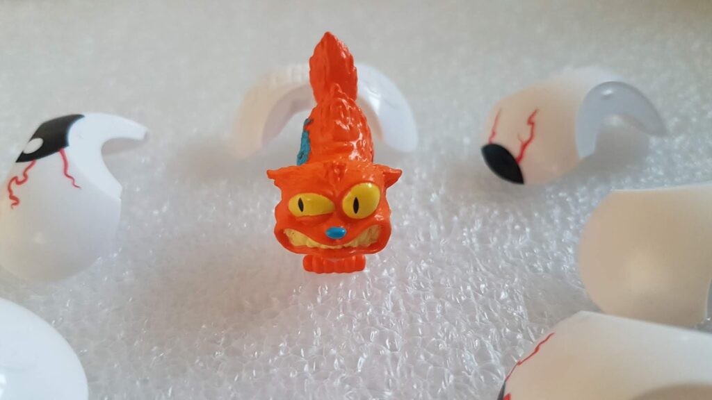 A small, orange, cartoonish cat figurine with exaggerated yellow eyes and a toothy grin stands on bubble wrap, surrounded by four broken white plastic pieces with painted black pupils and red veins resembling eyeballs.