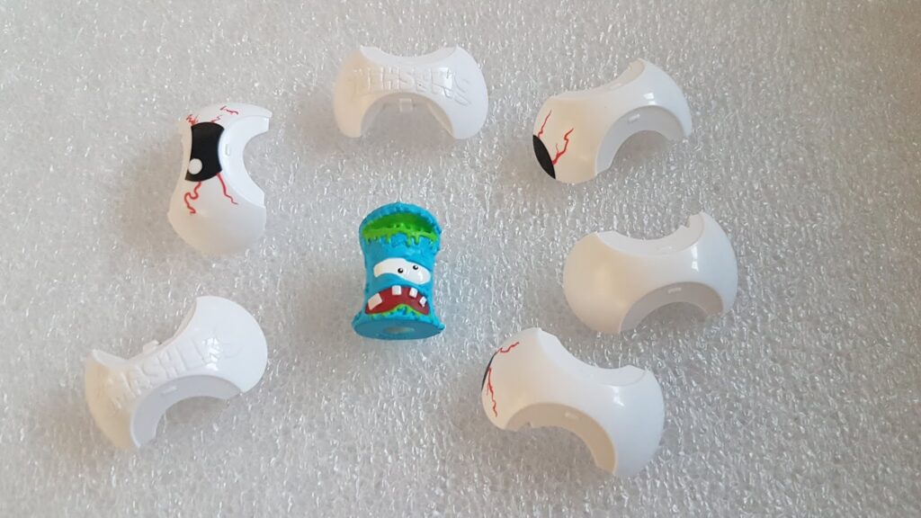 Six white, plastic, eyeball-shaped toy pieces with red veins surround a single blue, cylindrical toy figure with a face, sharp teeth, and a jagged green top. All toys are arranged on a textured white surface.
