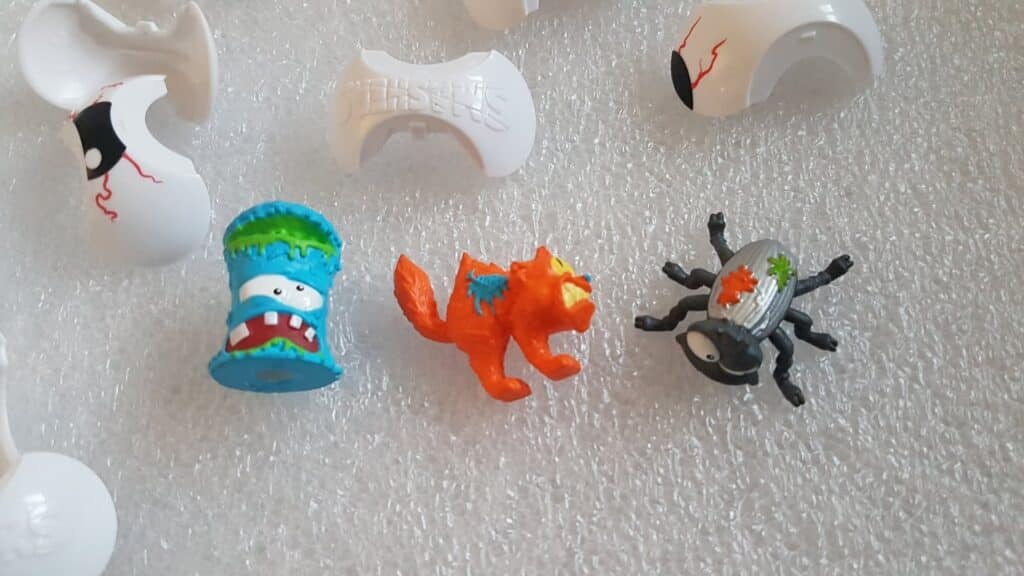 Three colourful toy figures are shown: a blue monster face in a cup, an orange cat with a fierce expression, and a grey-black spider with green and orange paint spots. Around them are broken white plastic shell pieces with red lines. The background is textured white.
