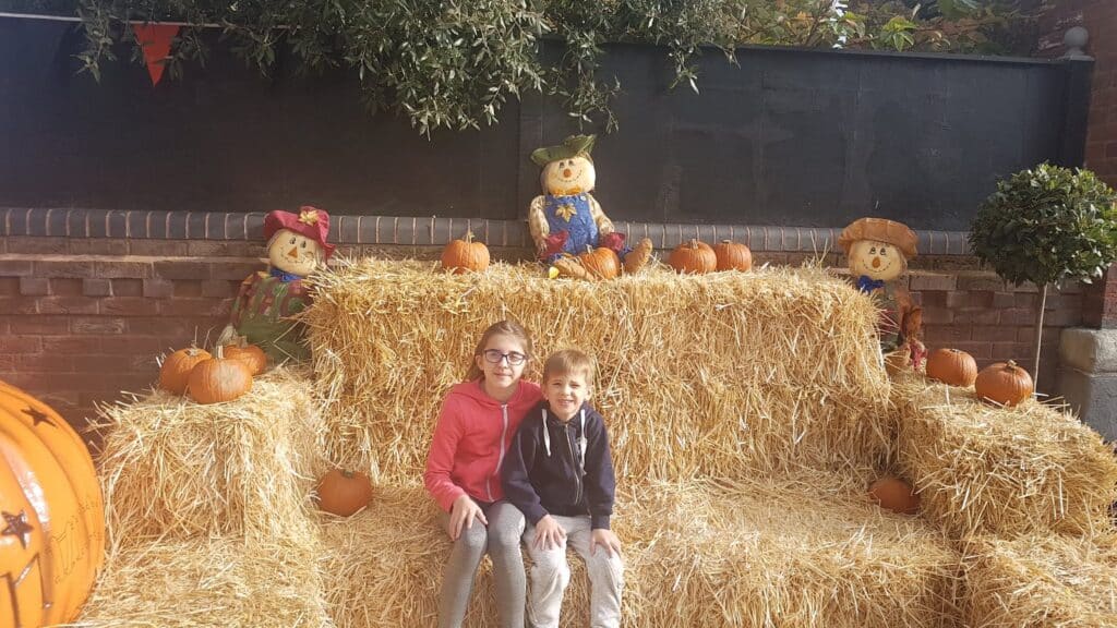 Two children sit on a large straw bale bench, surrounded by pumpkins. Three scarecrows and more pumpkins decorate the background. The setting appears festive and autumn-themed, with sunlight illuminating the scene.