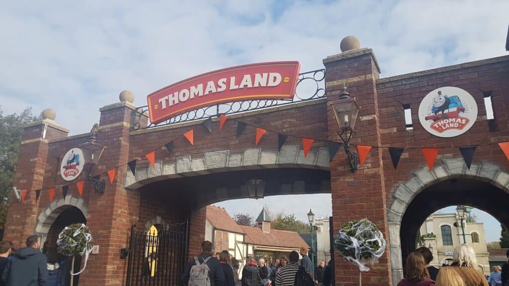 A brick entrance archway with a large red sign reading "Thomas Land" and smaller round signs with Thomas the Tank Engine. Black and orange bunting is strung across, and groups of people walk through under a partly cloudy sky. Lamp posts and cobweb decorations are visible.