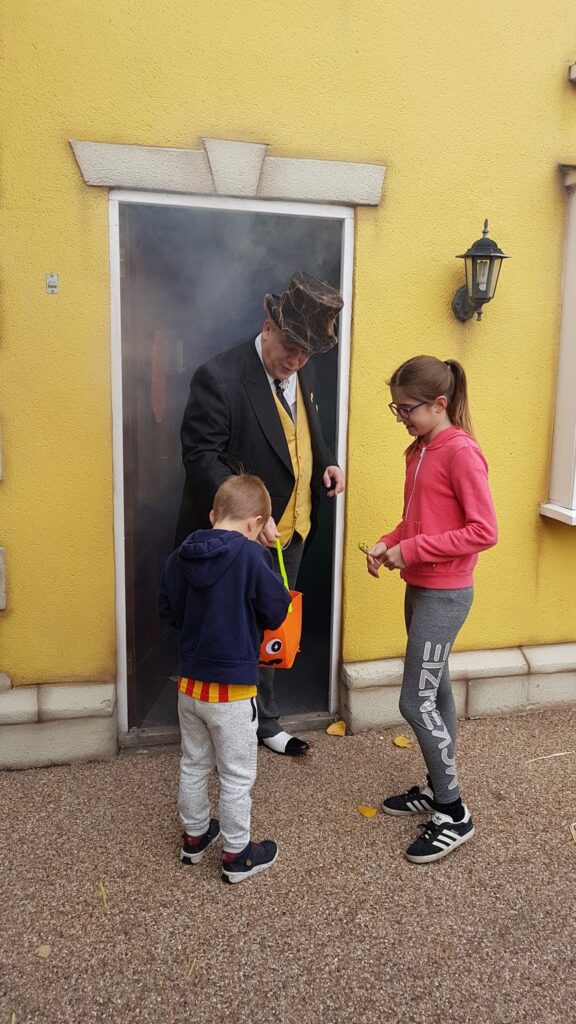 A man in a top hat and yellow waistcoat stands in a smoky doorway, handing out sweets to two children; one boy in a navy hoodie holding an orange bucket, and one girl in a pink hoodie and grey sweatpants, in front of a yellow wall with a black lantern.