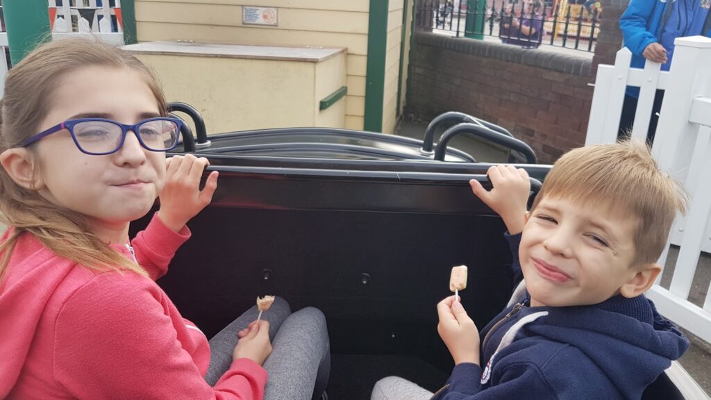 Two children, a girl in glasses and a pink hoodie and a boy in a blue jacket, sit in a ride car eating ice cream. Both are smiling at the camera. Behind them are a brick wall, a wooden building, and a white fence.