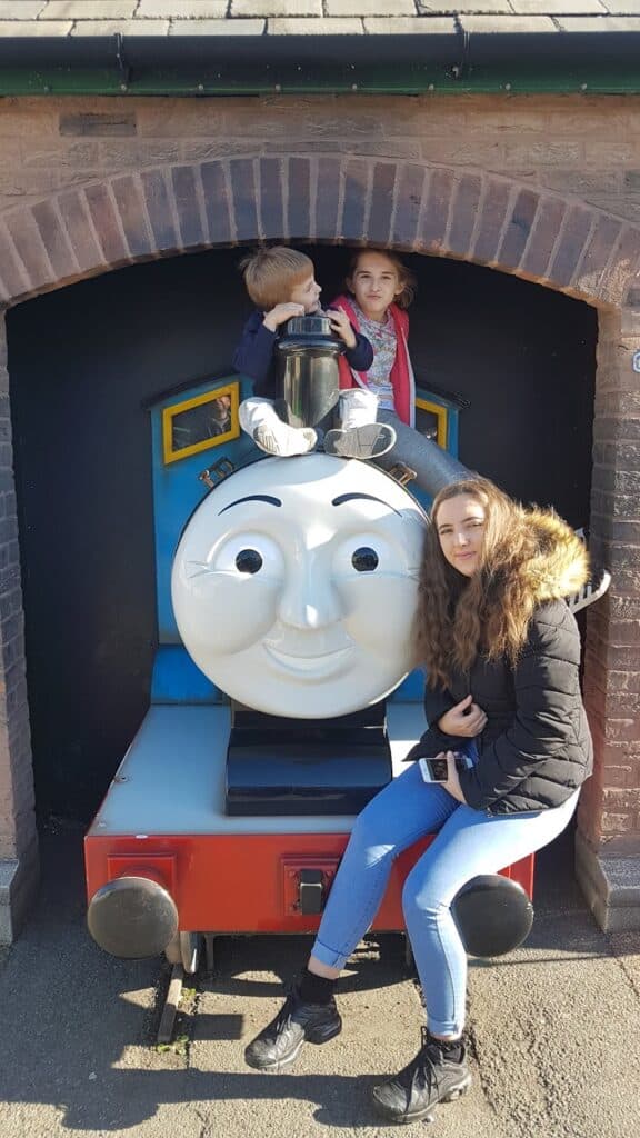Three children pose with a life-sized Thomas the Tank Engine model under a brick arch. Two sit on top, smiling, with one holding a toy train, while another sits on the front, wearing a black jacket and jeans, looking towards the camera.