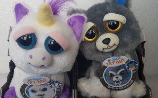Two soft toys are shown side by side. The left is a white unicorn with a yellow horn, purple ears, and big blue eyes. The right is a grey dog with floppy ears and big yellow eyes. Both have tags that say “Go Ahead… TRY ME! Squeeze the back of my head.”.