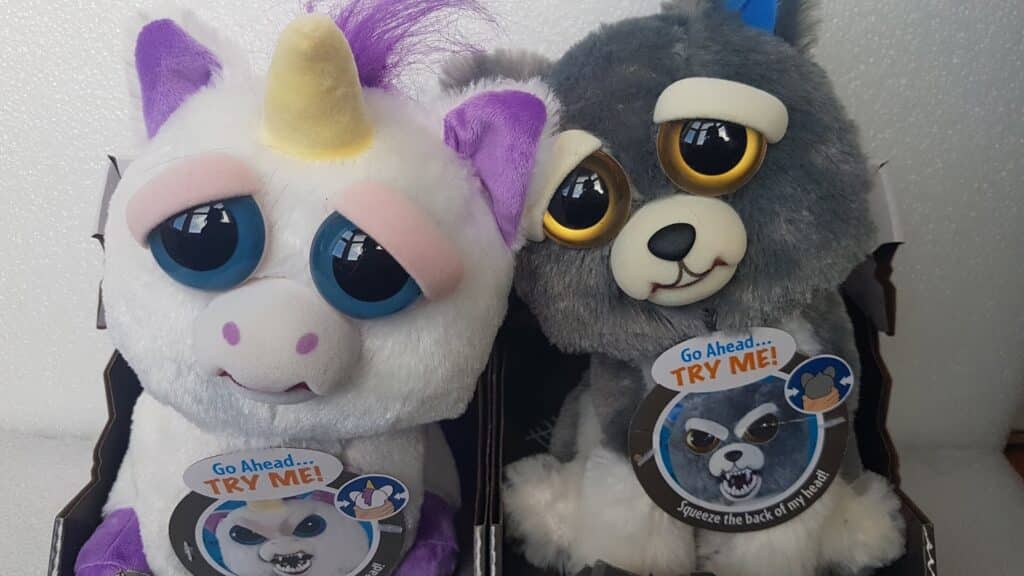 Two soft toys are pictured side by side: a white unicorn with a yellow horn and purple accents, and a grey wolf with big yellow eyes. Both have tags reading “Go Ahead… TRY ME!” with an image showing a fierce expression revealed when squeezed.