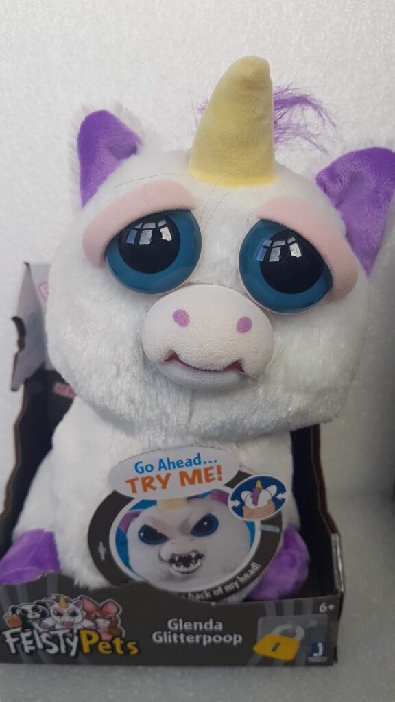 A stuffed unicorn toy with white fur, purple ears, a yellow horn, and big blue eyes sits in a display box labelled "Feisty Pets: Glenda Glitterpoop." A sticker says “Go Ahead… TRY ME!” inviting interaction to reveal a different expression.