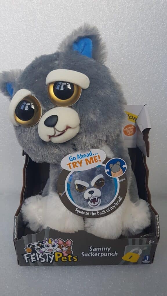 A plush grey and white wolf toy with large gold and black eyes is displayed in a Feisty Pets box. The tag reads, “Go Ahead… TRY ME! Squeeze the back of my head!” Its name, “Sammy Suckerpunch,” appears on the box. The background is plain.