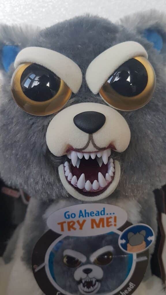 A plush toy wolf with grey fur, large round yellow eyes, white eyebrows, and an open mouth showing sharp white teeth. The toy looks angry or fierce. Attached is a tag with “Go Ahead… TRY ME!” and an illustration of the same wolf’s face.