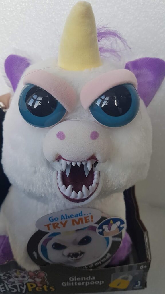 A plush unicorn toy with a white face, large blue eyes, a yellow horn, and purple ears. Its mouth is open showing sharp teeth in an aggressive expression. A tag reads "Go Ahead...TRY ME!" and the box says "Glenda Glitterpoop".