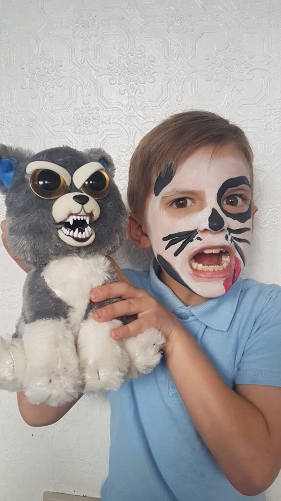 A child with face paint resembling a cat, wearing a blue shirt, poses making a fierce expression. They hold a grey and white plush cat toy with large eyes, bared teeth, and sunglasses in front of a textured white wall.