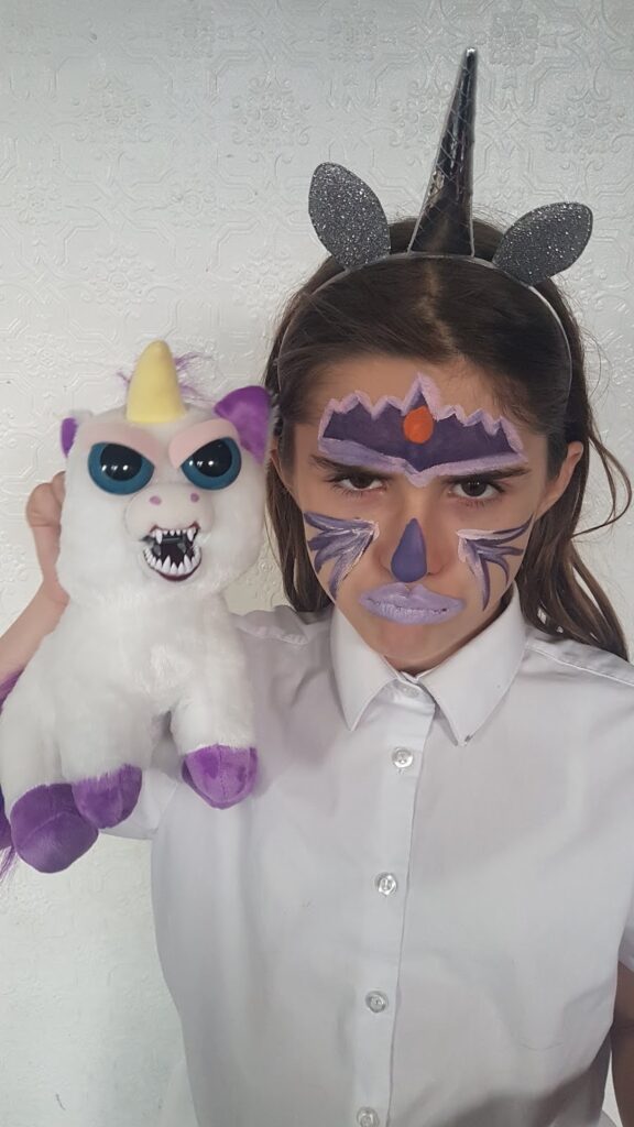 A girl dressed in a white shirt wears a unicorn horn headband with glittery ears and has elaborate purple and black face paint. She holds a white unicorn soft toy with big eyes, purple hooves, and an open mouth showing sharp teeth.
