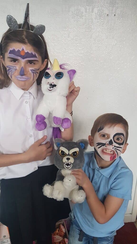 Two children with face paint hold soft toys. The girl has unicorn-themed make-up and wears a unicorn headband, holding a cuddly unicorn. The boy has an animal or monster face paint, wearing a light blue shirt, and holds a cuddly wolf. Both are smiling indoors.