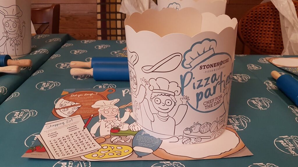 A paper chef’s hat with cartoon pizza-themed illustrations and the text “STONEHOUSE Pizza Party! CHEFS FOR THE DAY!” sits on a teal tablecloth. Nearby are rolling pins and a pizza activity sheet with a wordsearch and cartoon ingredients.