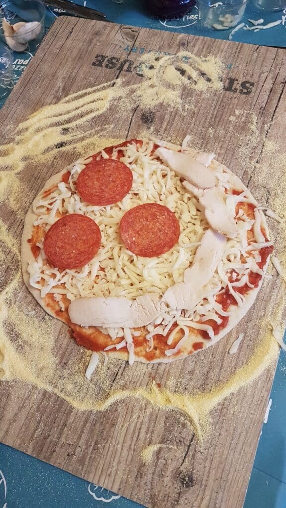 A homemade pizza on a wooden board topped with grated cheese, three slices of pepperoni, and several pieces of cooked chicken breast. Cornmeal is scattered on the board and under the pizza, and faint text is visible on the board.