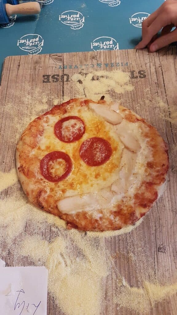 A small pizza on a flour-dusted wooden board, topped with melted cheese, three slices of pepperoni, and several slices of chicken forming a smiley face. A hand rests at the top right, and a blue surface with “Pasta Poetry” logos is visible in the background.