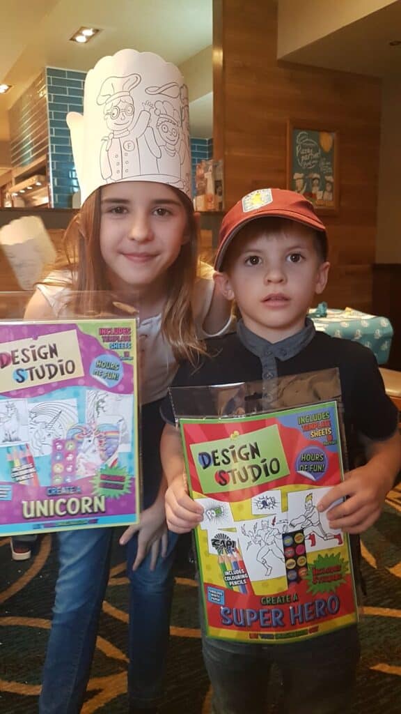 Two children stand indoors, smiling and holding “Design Studio” colouring kits. The girl wears a tall paper chef’s hat and holds a unicorn-themed kit. The boy wears a red cap and holds a superhero-themed kit. The background shows a restaurant with wooden decor.
