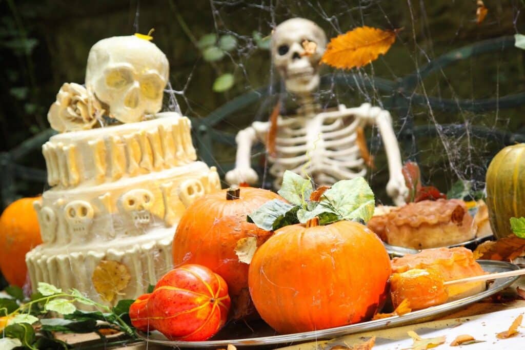 A Halloween-themed table displays pumpkins, a white skull-decorated cake, pie, and autumn leaves. In the background, a skeleton sits among cobwebs and more leaves, creating a spooky autumn atmosphere.