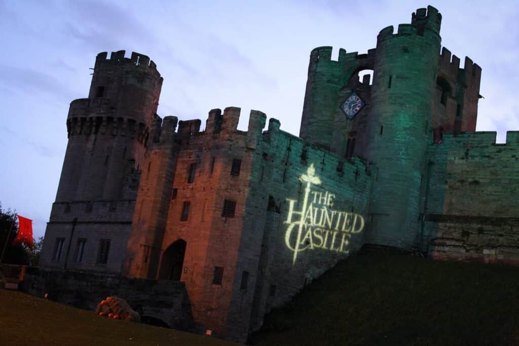 A large stone mediaeval castle at dusk, illuminated by green lighting. "THE HAUNTED CASTLE" is projected onto the walls in white text with a ghost graphic. The sky is darkening, and the foreground is grassy, adding to the eerie atmosphere.