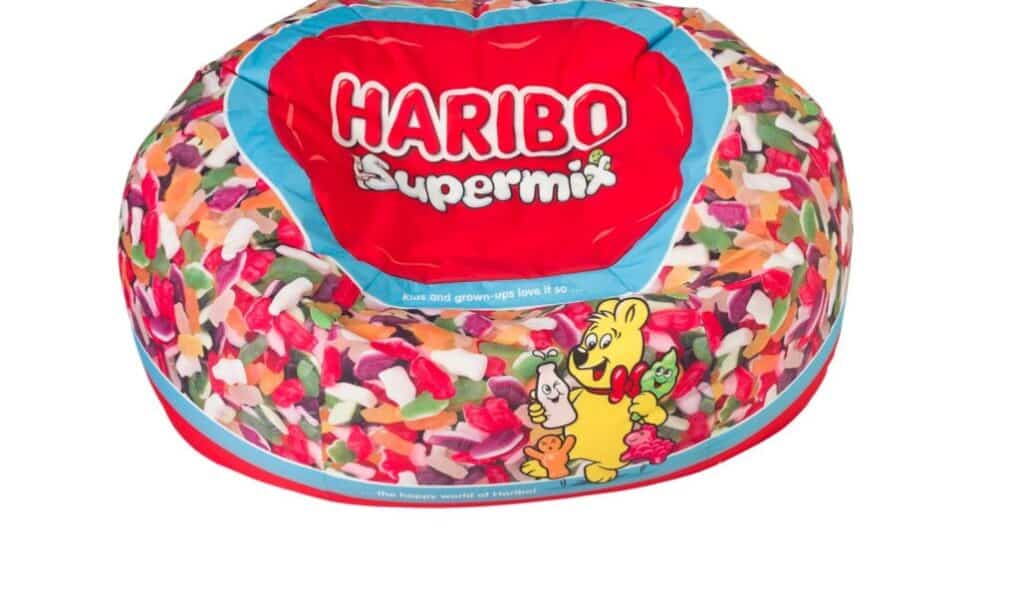 A colourful bean bag designed with images of assorted Haribo sweets, featuring a red area at the top with the white text "HARIBO Supermix" and a cartoon bear holding sweets on the side. The background is white.