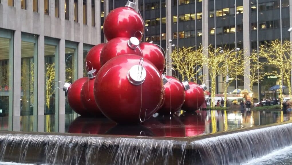Large, shiny red Christmas bauble sculptures stacked in a pyramid shape sit on a reflective black platform surrounded by water fountains. In the background are tall office buildings with yellow festive lights wrapped around tree trunks.