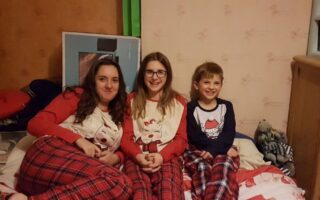 Three children sit on a bed, smiling at the camera. The two girls on the left wear matching red checked pyjama bottoms and cream tops with reindeer designs. The boy on the right wears red checked pyjama bottoms and a navy shirt. Soft toys are visible on the bed beside them.