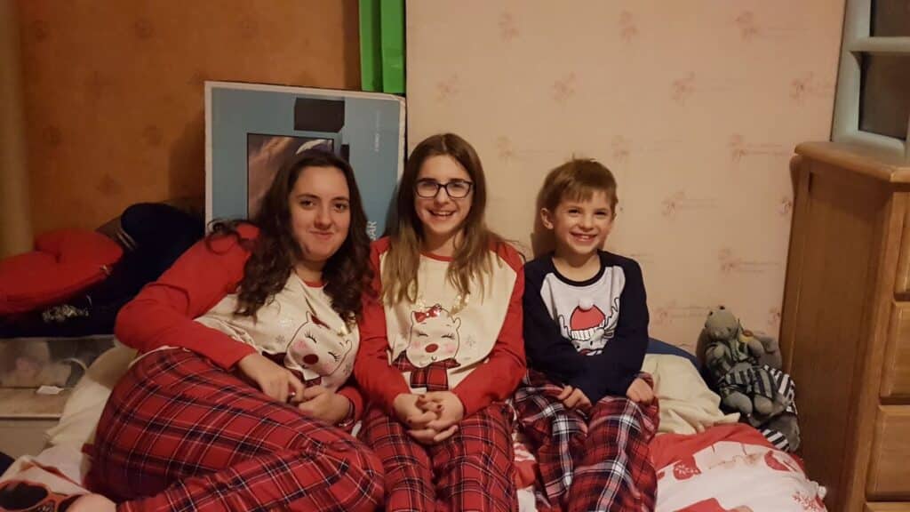 Three children sit on a bed wearing red checked pyjama bottoms. The two girls on the left wear matching cream and red tops with cat graphics; the boy on the right wears a navy shirt with a Father Christmas graphic. They smile at the camera, with toys and furniture in the background.