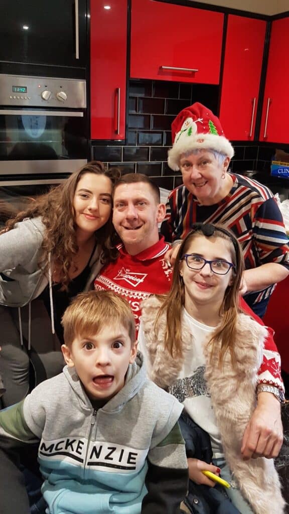 Five people, including two children and three adults, pose and smile in a kitchen with bright red cupboards. One woman wears a Santa hat. The boy in front pulls a wide-eyed funny face, adding a playful touch to the warm, festive group photo.