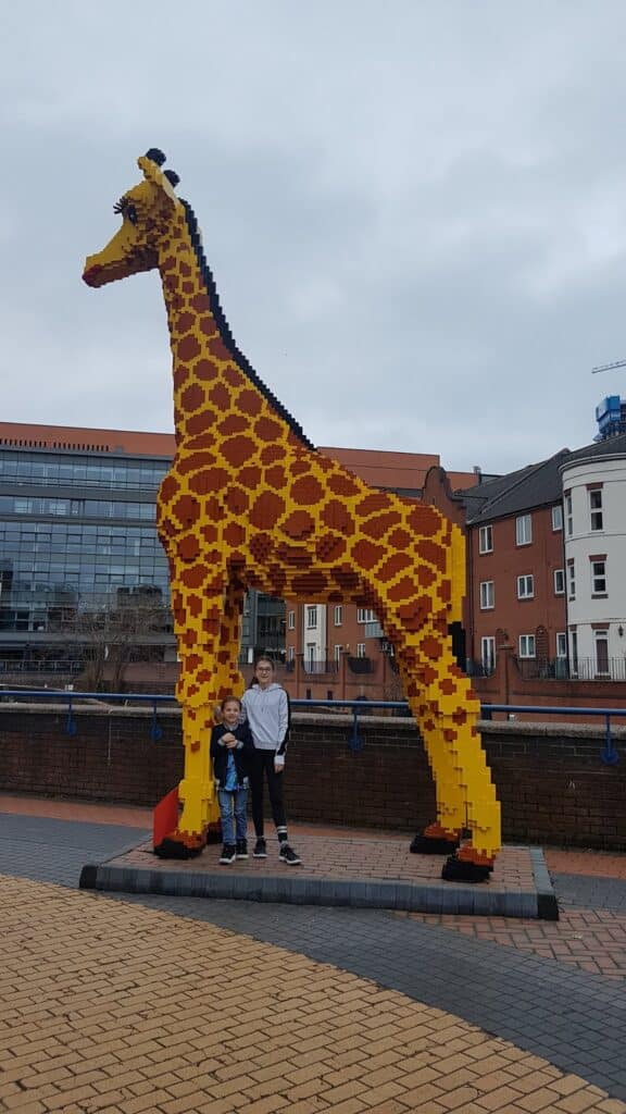 Two children stand in front of a large giraffe sculpture made from yellow and brown Lego bricks. The giraffe is outdoors on a paved area, with buildings and a cloudy sky in the background. The children are smiling and appear happy.