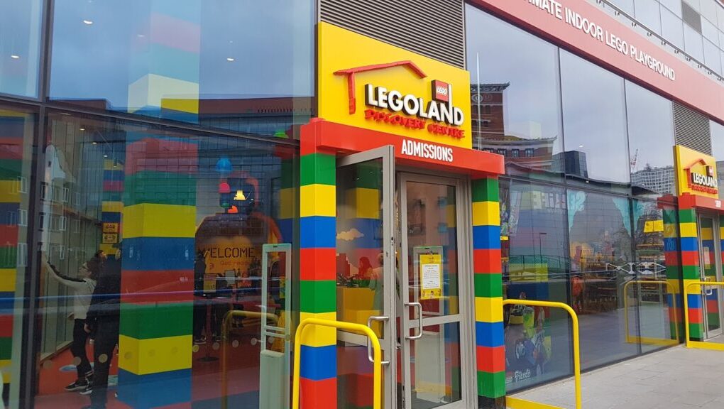 Entrance to LEGOLAND Discovery Centre with colourful LEGO brick-style walls, yellow railings, and a glass door labelled "ADMISSIONS." Reflections and visitors are visible through the glass. The adjacent building has red trim and large windows.