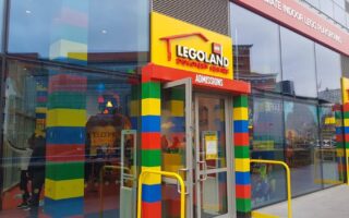 Entrance to LEGOLAND Discovery Centre with colourful LEGO brick-style walls, yellow railings, and a glass door labelled "ADMISSIONS." Reflections and visitors are visible through the glass. The adjacent building has red trim and large windows.