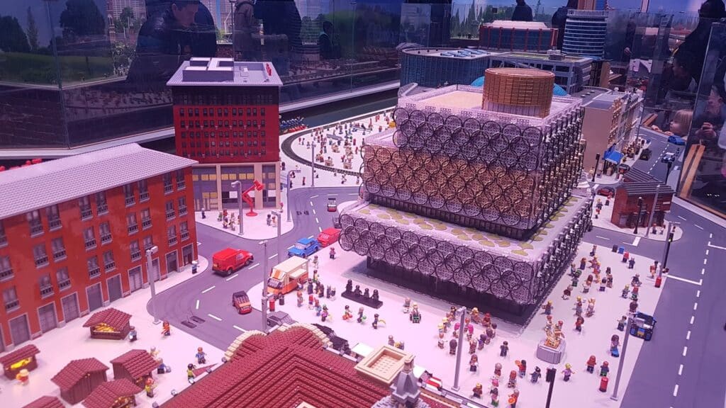 A large, detailed LEGO city features many minifigures, roads, cars, and buildings. A prominent modern building with a circular-patterned façade sits at the centre. Other red-brick structures and numerous tiny figures fill the lively urban scene.