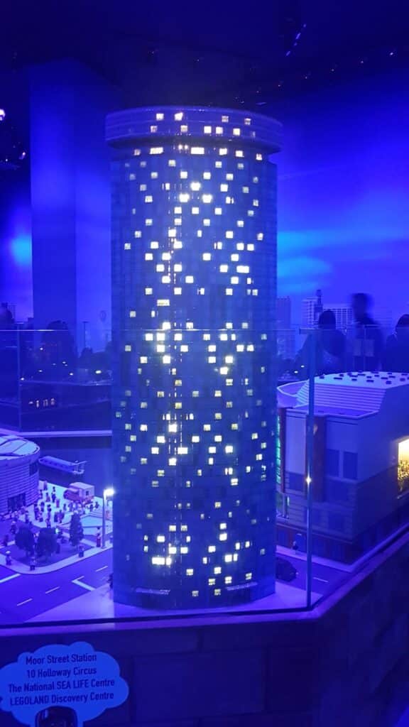 A tall, cylindrical LEGO building glows with blue and yellow lights in a nighttime city scene. Miniature cars, buses, and other buildings surround it. A sign in the foreground lists attractions: Moor Street Station, National SEA LIFE Centre, and LEGOLAND Discovery Centre.