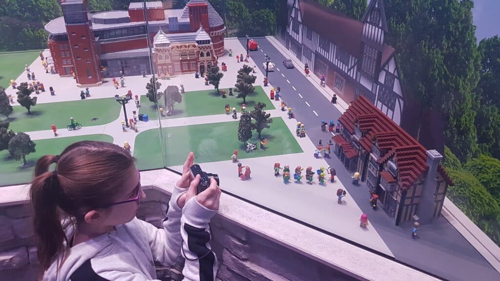 A young girl with glasses and a ponytail, wearing a striped jacket, holds a camera and looks at a large, detailed model city behind glass. The model features buildings, trees, cars, and many small figurines on streets and pavements.