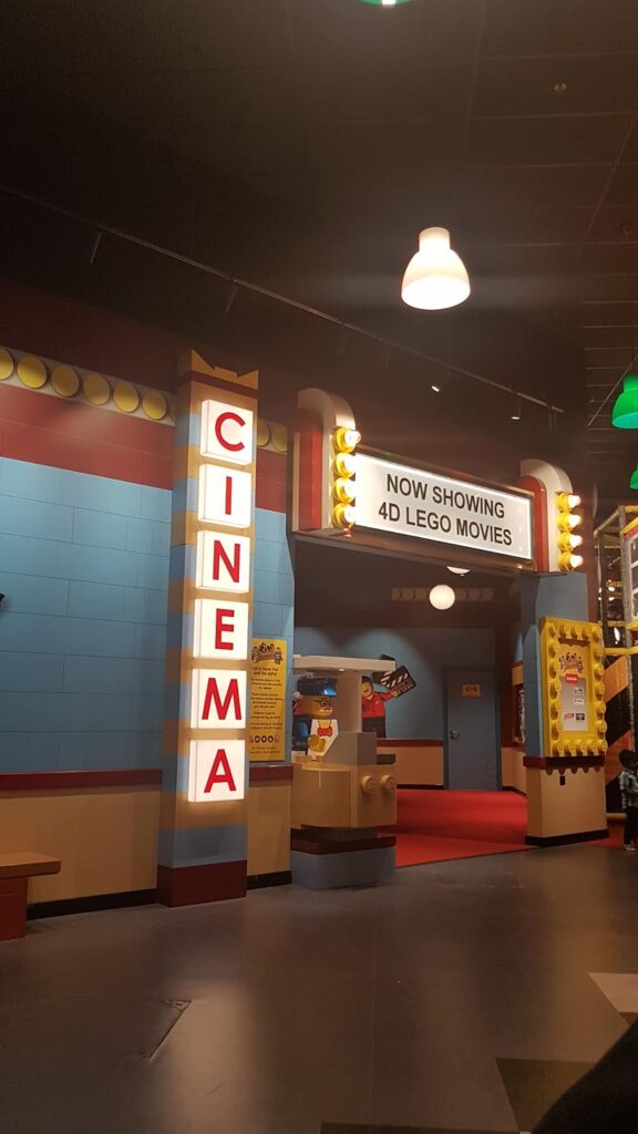 Indoor scene of a LEGO-themed cinema with bright sign reading “CINEMA” in vertical red letters and marquee announcing “NOW SHOWING 4D LEGO FILMS.” Popcorn stall and playful, colourful brick decor evoke a fun, child-friendly atmosphere.