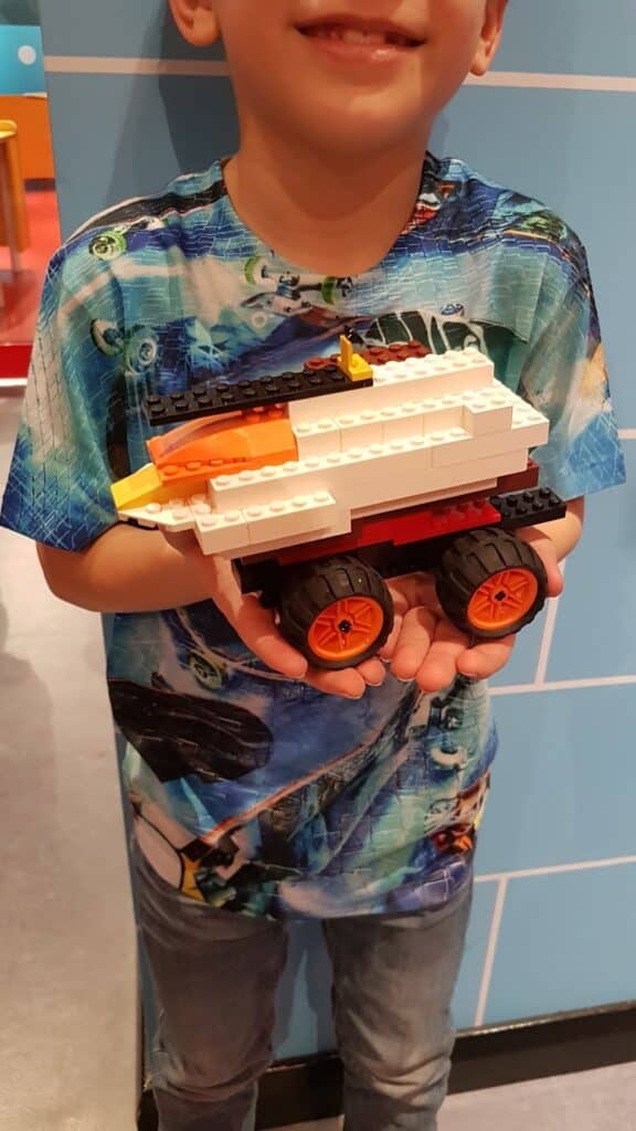 A child wearing a colourful shirt with cars holds a LEGO vehicle made of white, red, and orange bricks with four large wheels. The child is smiling, but only the lower half of their face is visible. The background shows a blue wall.